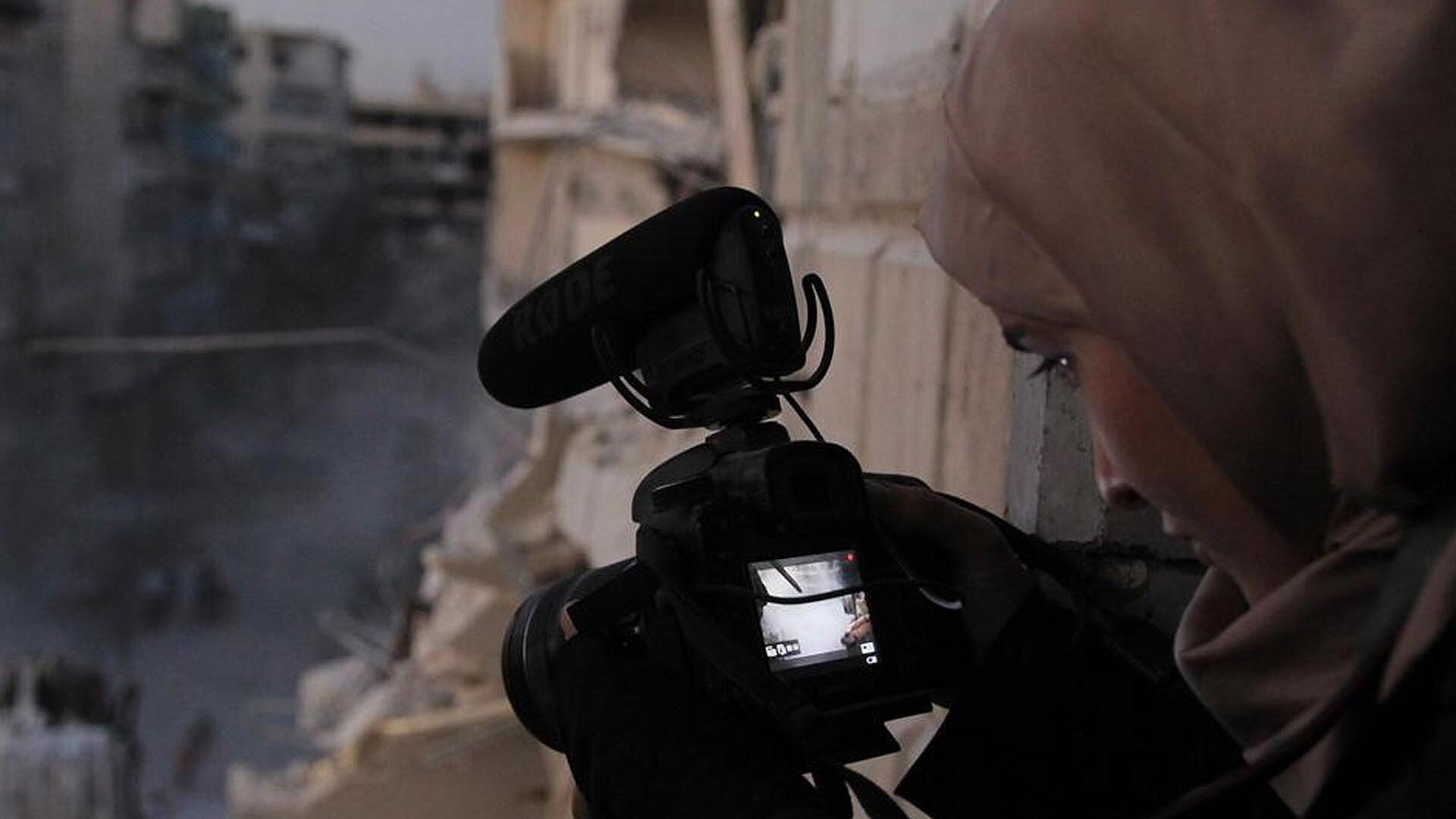 [Translate to Deutsch:] A young women holds a camera in a war torn town.